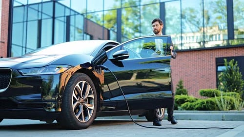 Geschäftsmann überprüft das Telefon, sein Elektroauto wird draußen an einer modernen Station aufgeladen