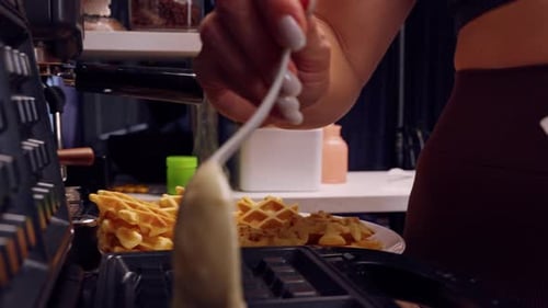 Side View Of Waffle Batter Dropping Onto Hot Iron Plates With Cooked Waffles In Background, Home