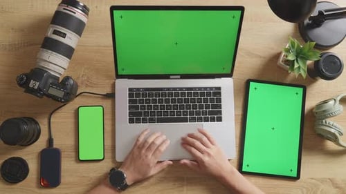 Vista superior de las manos de un editor de video masculino usando una computadora portátil de pantalla verde con un teléfono inteligente de pantalla verde