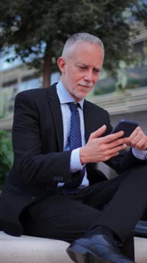 Vertical Smiling Mature Businessman with Suit Using a Smartphone Sitting Outside Relaxing and