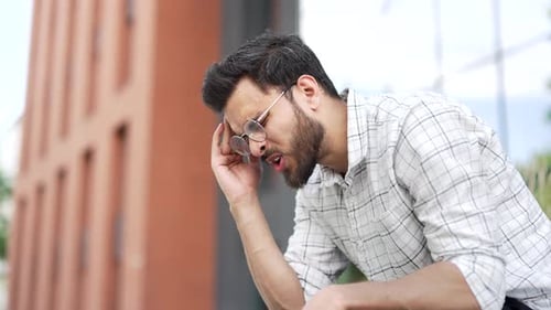 A young adult man is suffering from a headache while sitting on a bench on the street. Upset busines