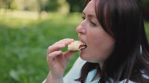 A brown-haired girl in a striped shirt sits on the grass in the park in the open air and eats pizza