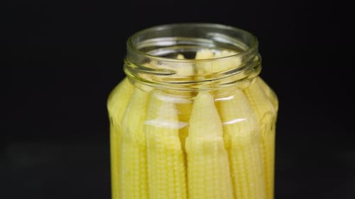 Marinated baby corn cobs in glass jar without lid rotating on black plate close up. Canned baby
