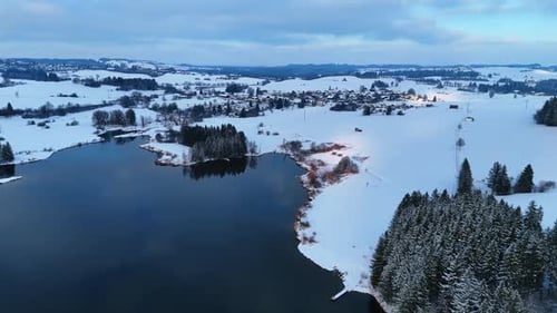 Footage over the river not frozen in winter. White snow covers the waterfront and pine trees.