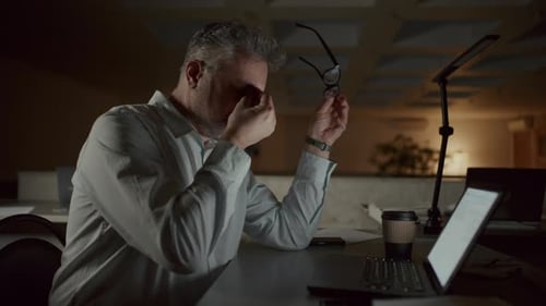 Idoso exausto esfregando os olhos enquanto trabalhava em um laptop até tarde no escritório