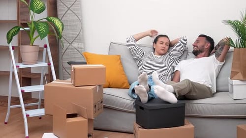Room Filled with Moving Boxes Tired but Happy Couple Relaxes on Sofa Discussing House Decoration