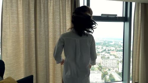 Woman in oversized shirt opens curtains to city view