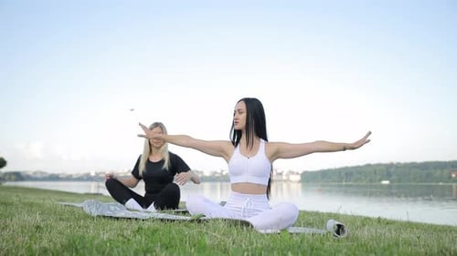 Two European Young Women are Doing Yoga While Sitting on the Shore of the Lake