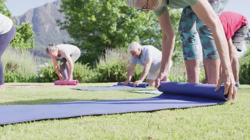 Diverse group of male and female seniors rolling up yoga mats after exercise in garden, slow motion