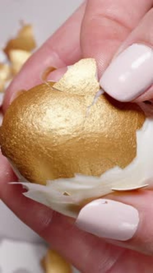 Vertical video. Detailed shot of hands peeling a golden-painted hard-boiled Easter egg, with shiny