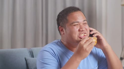 Close Up Of A Fat Asian Man Talking On Smartphone While Eating Fast Food Having Hamburger On A Sofa