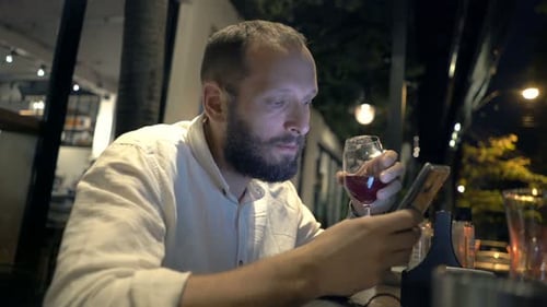 Young man enjoys red wine texting on smartphone at cafe counter at night