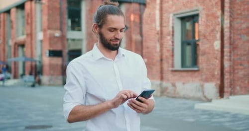 Portrait of handsome smiling man typing in mobile while looking into distance in urban street