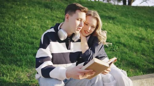 Joven pareja disfrutando de una tarde soleada mientras leen un libro juntos en una colina cubierta de hierba en un parque
