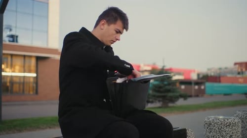 Man Seated Outdoors Going Through Briefcase Taking Out Tablet with Urban Building Background