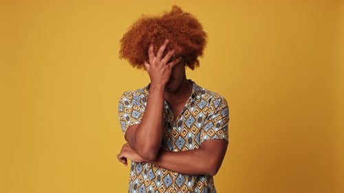 Bored guy is annoyed, covers his face with his palm isolated on yellow background in studio