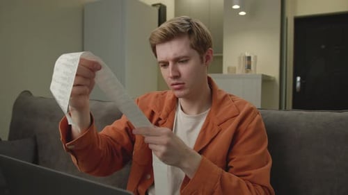 Erschöpfter Mann mit Quittung Junger erwachsener Mann ist verärgert über riesige Ausgaben, wenn er zu Hause auf der Couch sitzt