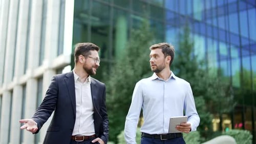 Two businessmen walk outside in front of an office building, talking and discussing work matters,
