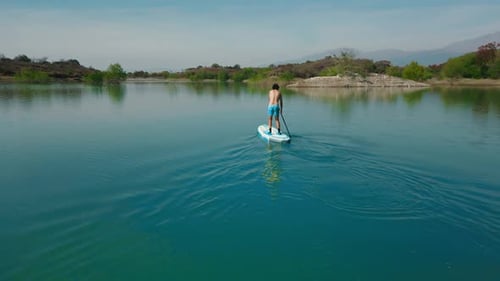 Aerial drone flying over a man doing stand up paddle in a blue water lake with mountains