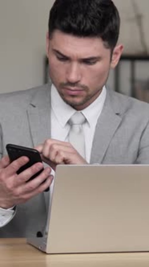 Man in Suit Uses Phone and Laptop at Desk