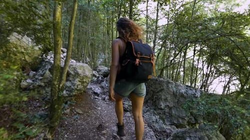 behind view of a young woman hiking walking in nature on a trail underneath green trees surrounded b