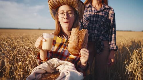 Young Girl Farmer in Sun Hat Eats Loaf of Bread Biting It Drinking Milk Sits on Wheat Field with