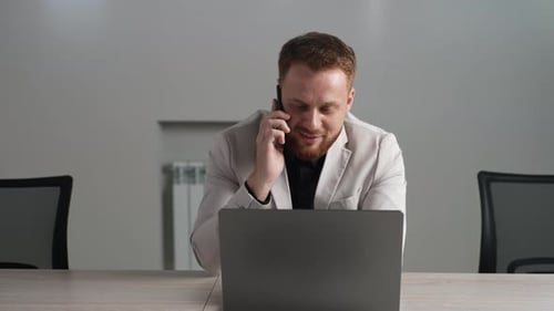 Portrait of Smiling Businessman in Suit Using Laptop Talking on Smartphone Sitting at Office Desk