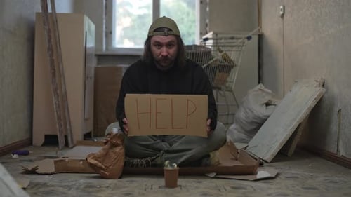 Homeless Poor Man Sitting in a Room of an Abandoned Building with a Money Jar He is Begging for Help