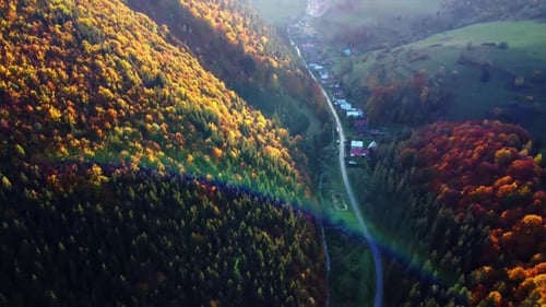 Aerial view of autumn valley with forested hills, winding road and sunlit rural village in foothills