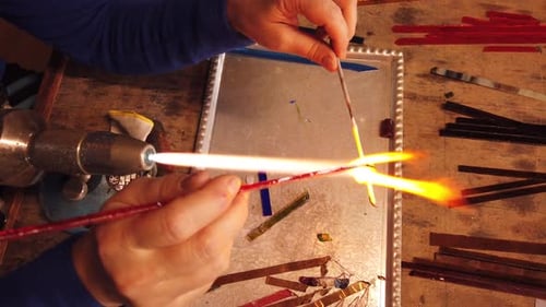 Plan rapproché de mains façonnant une perle de verre en la tenant au-dessus d'une torche à gaz dans un atelier. Bijoux en verre