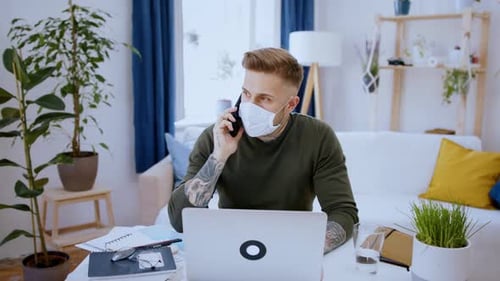 Man in home office works at desk with laptop smartphone and face mask