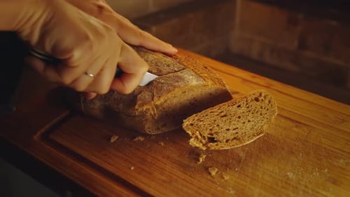 Close-up hands of female baker with knife cutting freshly baked aromatic loaf of wholemeal bread