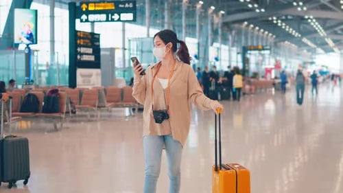 Woman walking with suitcase and camera while using smartphone at Airport Terminal