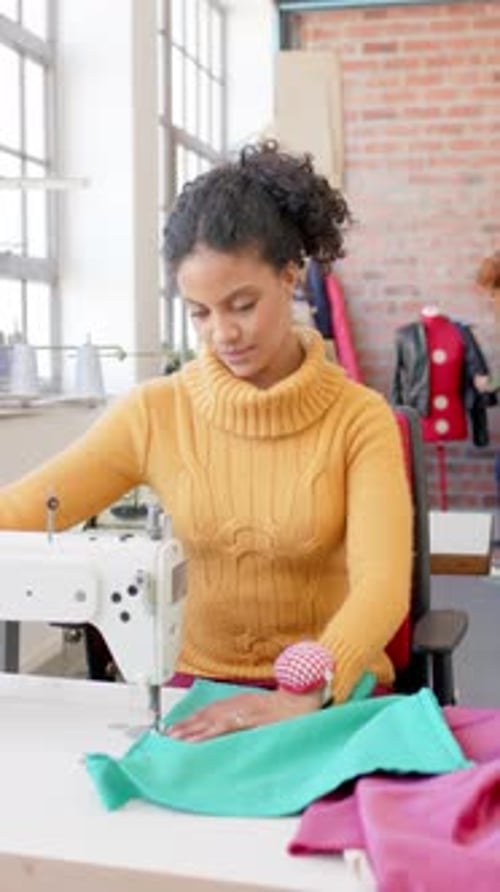 Diverse female fashion designer sewing fabric with sewing machine in studio, slow motion, vertical