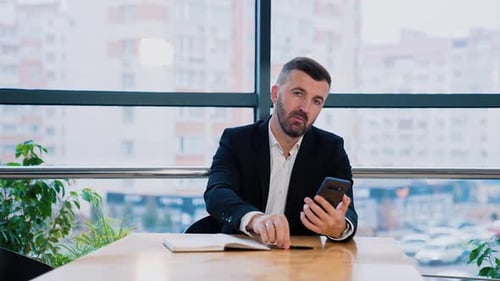 BBearded man in suit sits at the desk in his office. Businessman is looking at his phone