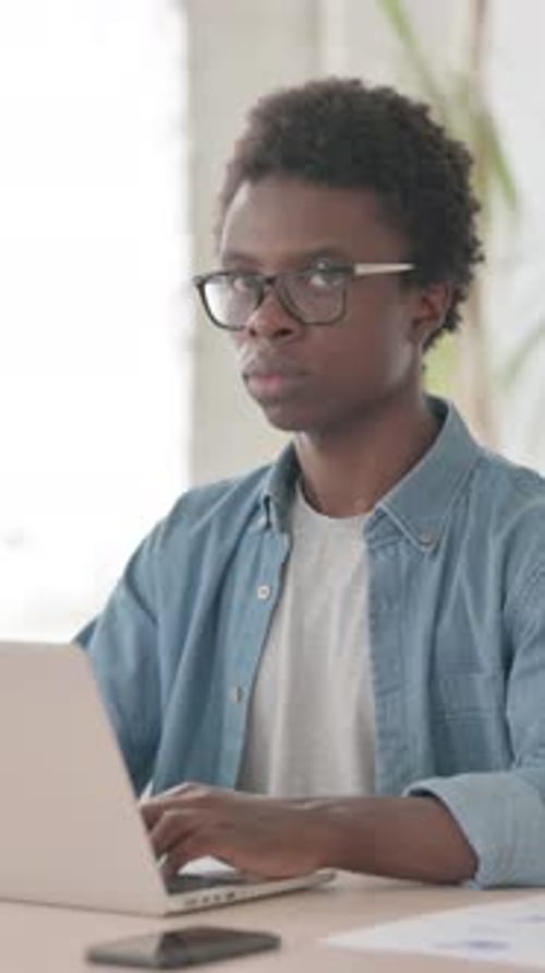 Young African Man Looking at Camera while using Laptop in Office, Vertical Video