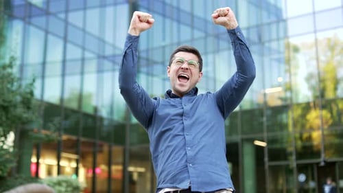 Happy excited businessman celebrating success and victory standing on street near modern office