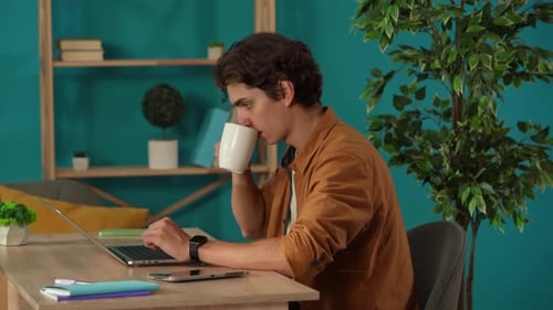 Medium Shot Capturing a Man Working on a Laptop in His Home Office and Drinking Coffee Young Male