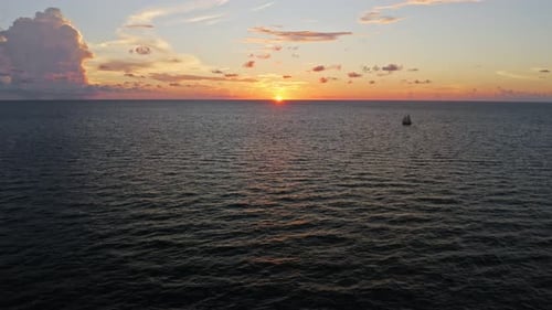 Sailboat silhouetted against fiery sunset on ocean horizon — aerial perspective