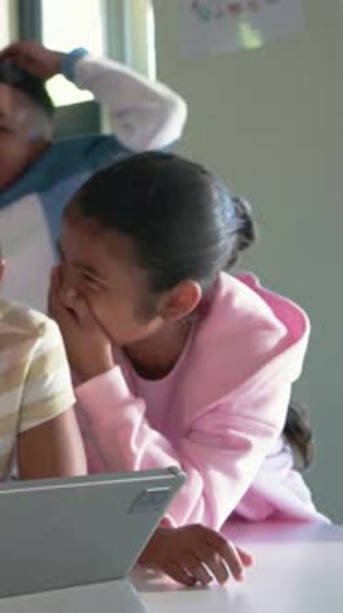 Vertical video: Leaning girl in pink hoodie watching clip on tablet at classroom window, reacting