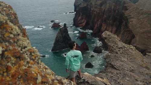 A man stands on the edge of a rocky cliff, overlooking the jagged volcanic coast and turquoise sea b