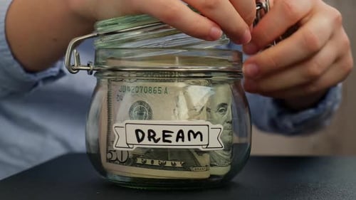 Female Hands Add Money to Glass Jar Full of American Currency Dollars Cash Banknote with Text DREAM