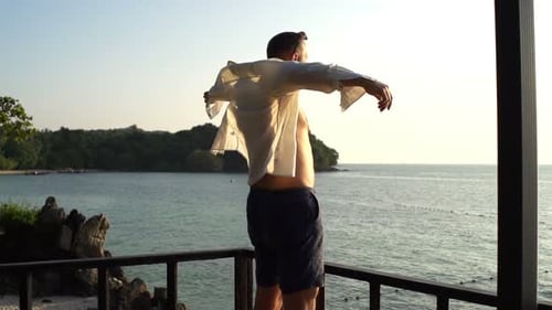Young man puts on shirt on terrace enjoying amazing seaside sunset in summer