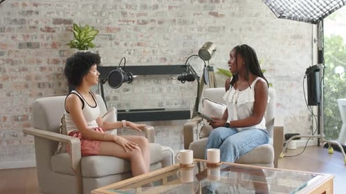 African American women host, guest hosting studio podcast using mics, host asking guest lifting mug