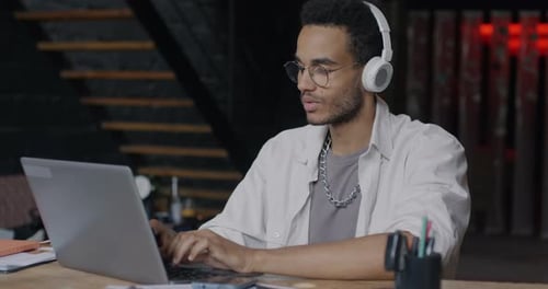 Carefree Biracial Office Worker Listening to Music Through Headphones and Using Laptop in Creative