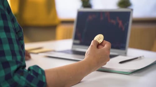 Adult Holding Bitcoin Coin Looking at Stock Graph
