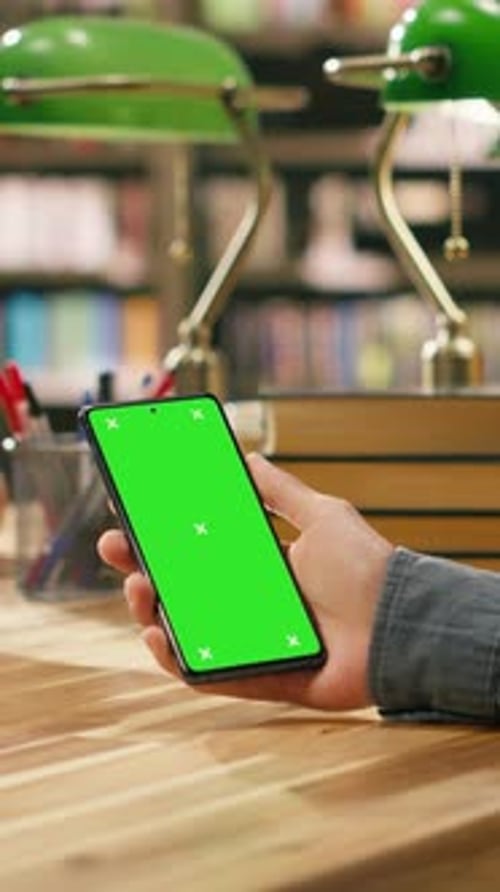 Vertikales Video Vertikaler isolierter Greenscreen auf einem Smartphone, das über einem hölzernen Bibliothekstisch gehalten wird