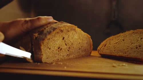 Close-up hands of female baker with knife cutting freshly baked aromatic loaf of wholemeal bread