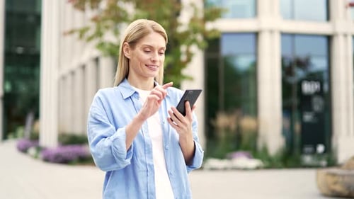 Happy blonde woman is using the phone while walking on the street near a modern building.