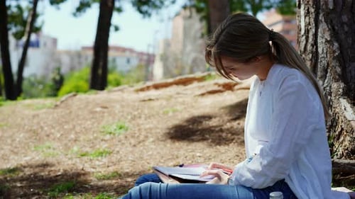 Focused Student Writing in Notebook Sitting Under Shady Tree Surrounded By Verdant Park Landscape